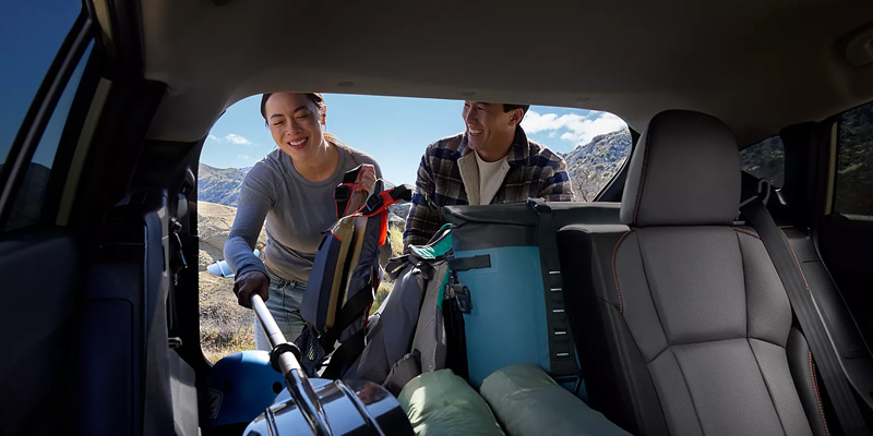 A couple loading camping gear into a Subaru car, surrounded by mountains and clear skies.