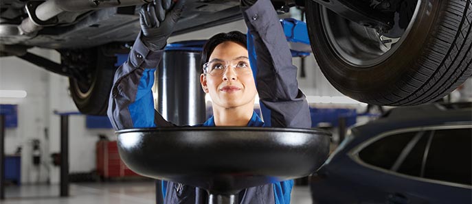 Female mechanic draining oil from a vehicle in a service garage, wearing protective glasses and work attire.