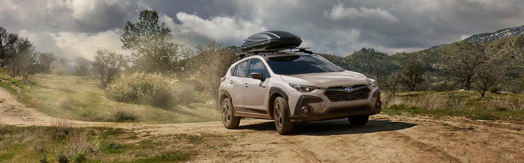 2026 Subaru Crosstrek driving on a dirt road with roof cargo carrier and scenic mountain landscape under cloudy skies.