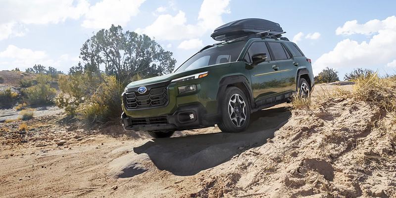 2026 Subaru Outback in green driving down a sandy off road trail with roof cargo box and rugged terrain background.